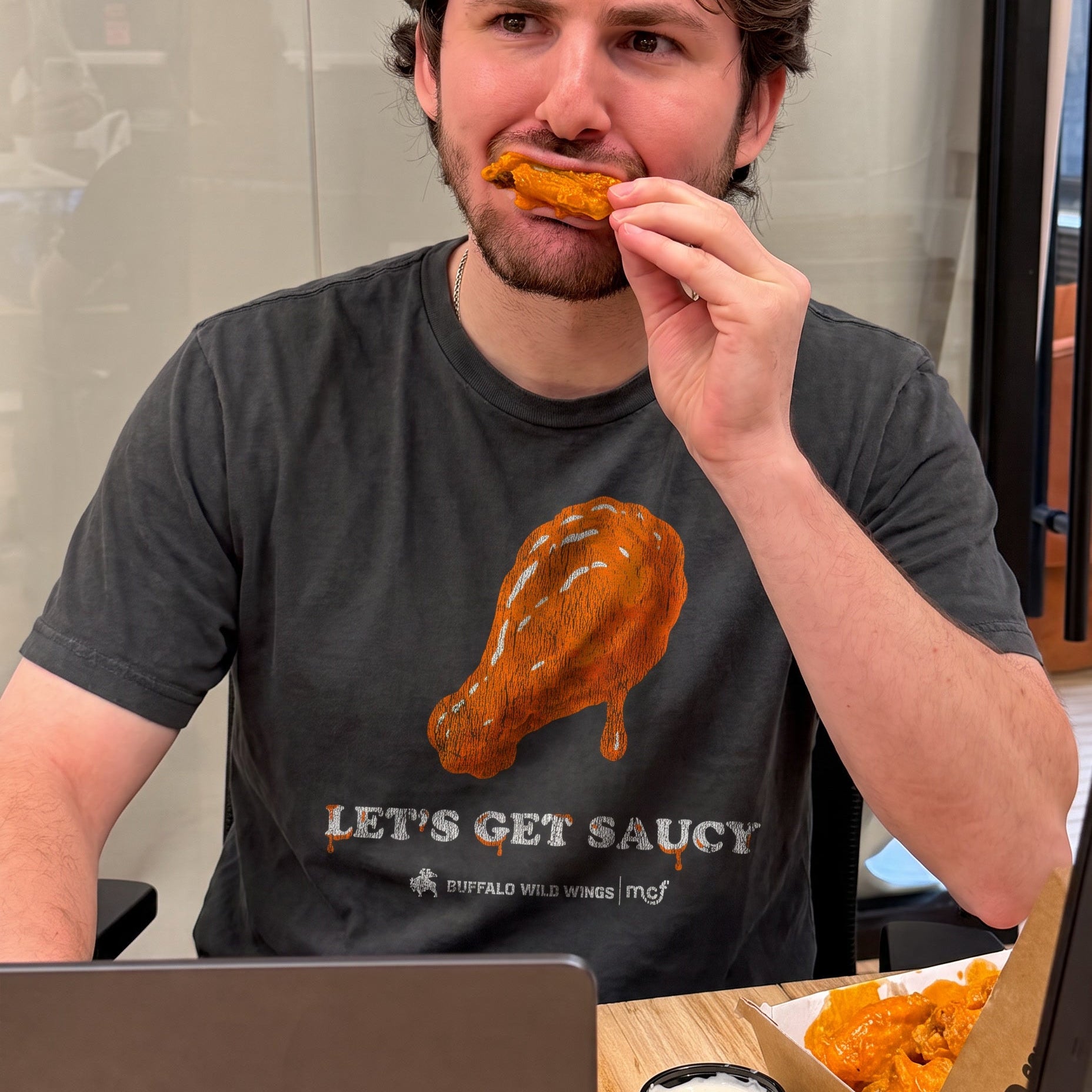 A man wearing a dark gray t-shirt with a graphic of a chicken wing in orange-brown sauce and the text 'LET'S GET SAUCY' below it, eating a piece of chicken wing at a restaurant table with a laptop, dipping sauce, and a box of wings.