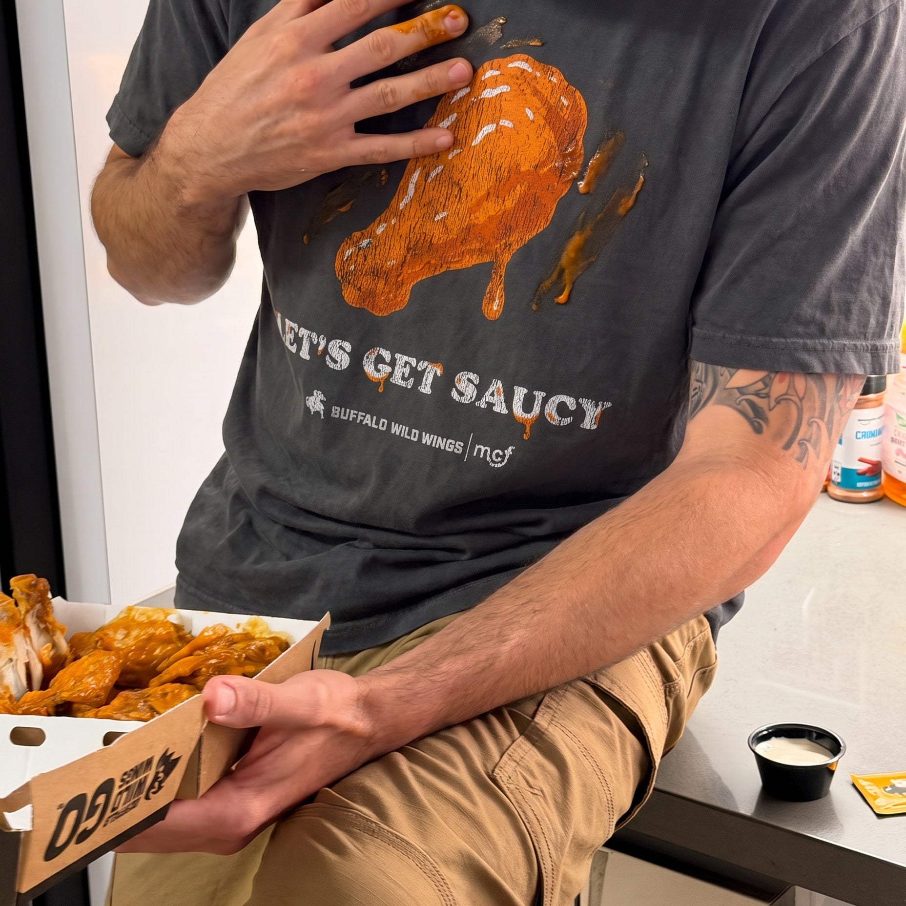 A man wearing a dark gray t-shirt with a graphic of a chicken leg covered in orange sauce and the text 'LET'S GET SAUCY' and 'BUFFALO WILD WINGS | mad', holding a box of Buffalo Wild Wings wings.
