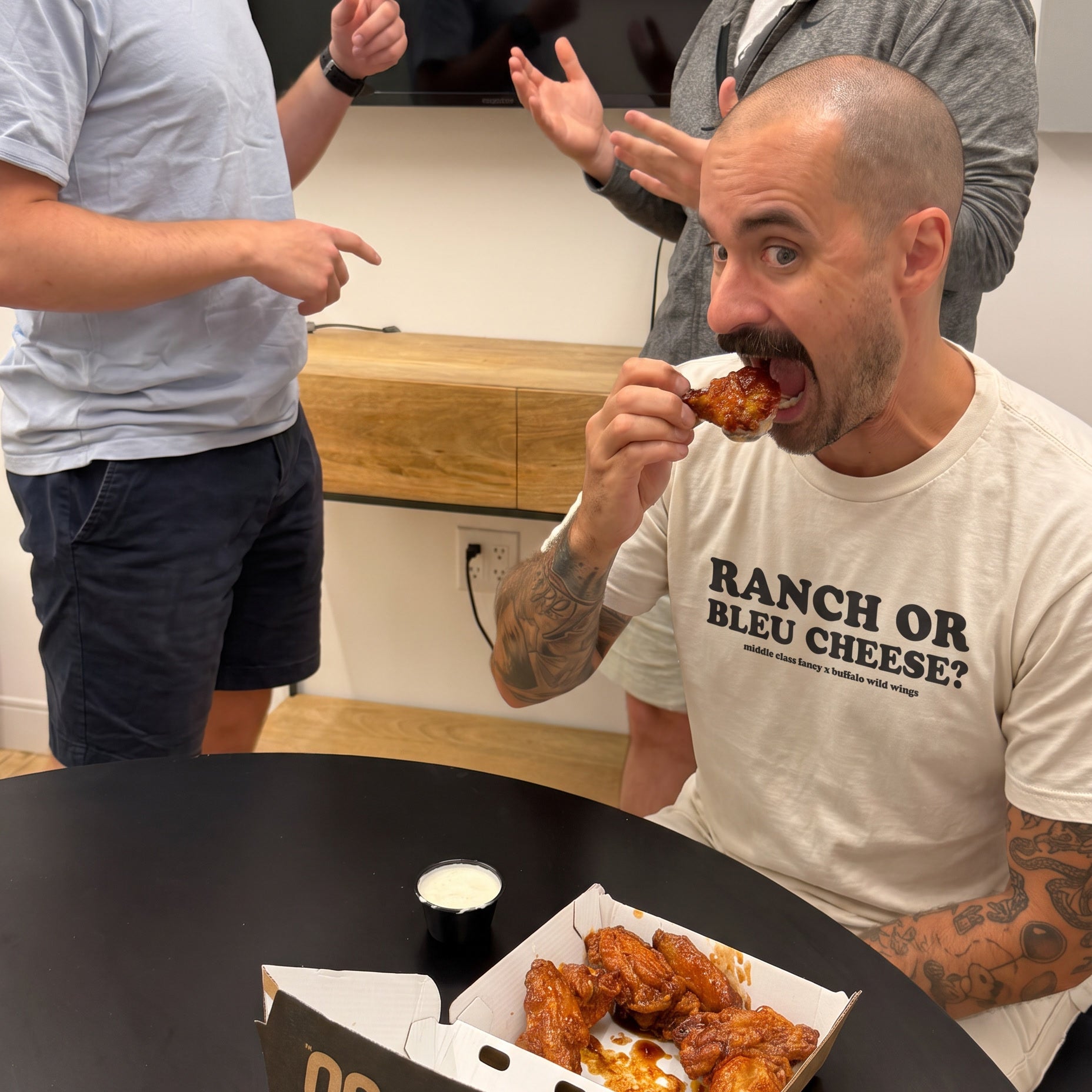 A man in a white graphic t-shirt eating chicken wings from a box labeled 'Ranch or Bleu Cheese' at a black table, with two other men gesturing in the background.